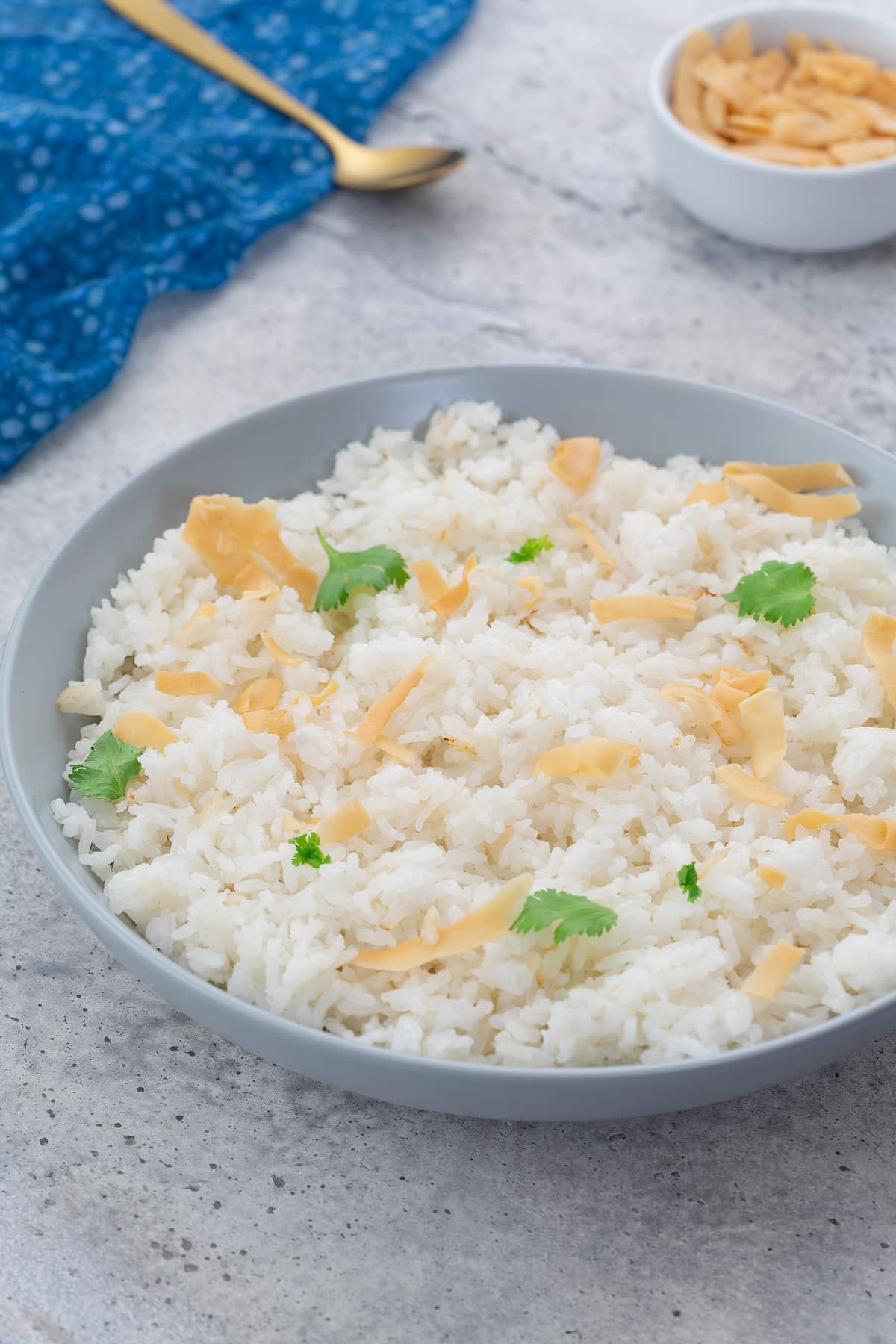 Coconut rice in a white bowl, garnished with coconut flakes and cilantro, placed on a white table. A blue towel, golden spoon, and a cup of coconut flakes are arranged nearby.