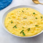 Creamy homemade polenta in a white bowl on a white table, with a golden spoon and a blue towel nearby.