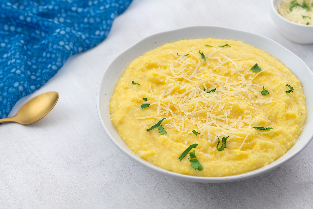 Creamy homemade polenta in a white bowl on a white table, with a small cup of cheese, a golden spoon, and a blue towel placed nearby.