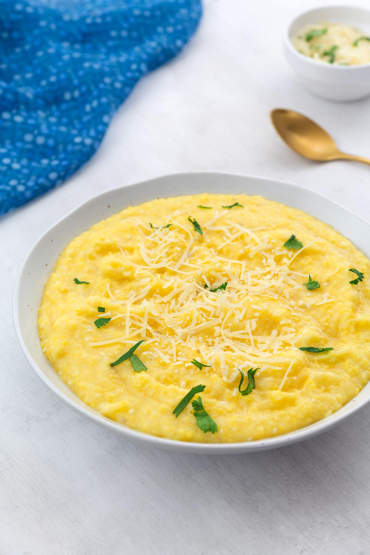 Creamy homemade polenta in a white bowl on a white table, with a small cup of cheese, a golden spoon, and a blue towel placed nearby.