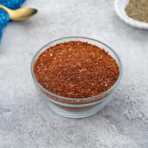 Homemade dry rub for ribs in a glass bowl on a white table, with a small plate of black pepper, a golden spoon, and a blue towel in the top corners.