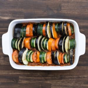 A baking dish with vegetables layered over tomato sauce, seasoned with spices.