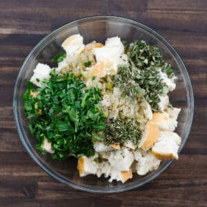 A bowl with crusty bread cubes, sautéed aromatics, and fresh herbs, ready to be mixed.