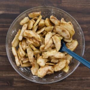 Apple slices tossed in sugar and spices in a bowl, with a blue spatula resting inside.