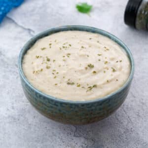 A bowl of cream of chicken soup served in a patterned green bowl on a white table, surrounded by a small spice container with dried herbs, a plate of pepper powder, a blue towel, and a decorative leaf.
