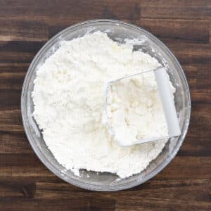 A glass bowl of flour crumbled with fats using a pastry cutter.