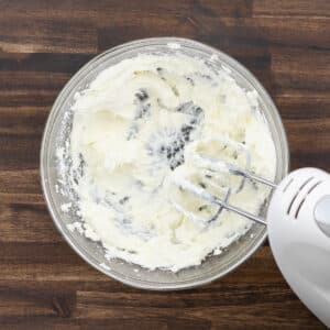 A bowl of cream cheese and powdered sugar blended into a smooth mixture, with an electric beater placed beside it.