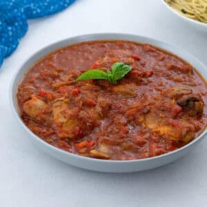 Homemade Chicken Cacciatore served in a bowl on a white table, accompanied by a blue towel and a bowl of aglio e olio.