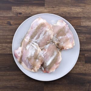 A plate with raw chicken thighs seasoned with salt and black pepper, ready for cooking.