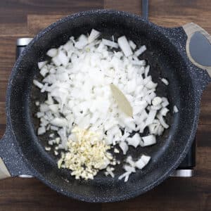 A pan with chopped onions, minced garlic, and a bay leaf, ready for sautéing.
