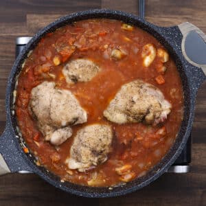 A pan with seared chicken thighs nestled in a tomato-based sauce, beginning to simmer.