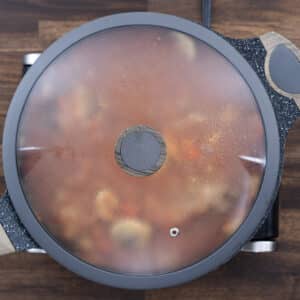A pan with chicken thighs simmering in a flavorful tomato sauce, covered with a lid.