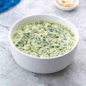 A bowl of creamy spinach on a white table, accompanied by a small plate of garlic cloves and a blue towel.