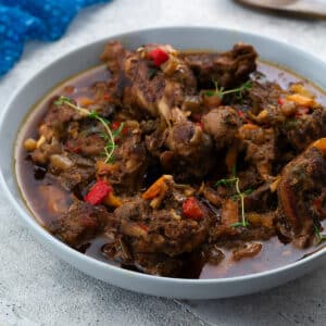Jamaican Brown Stew Chicken in a light gray bowl on a white table, with a blue towel and wooden spoon placed in the top corners.