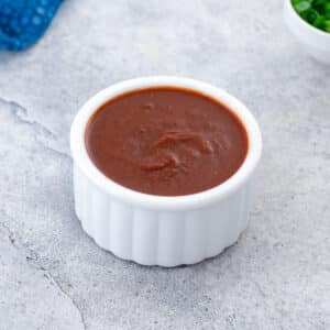 Homemade taco sauce in a white bowl on a white table, with a blue towel and chopped green onions placed in the top corners.