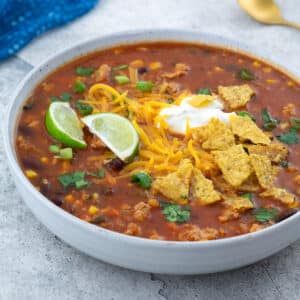 A white bowl of homemade taco soup topped with cheddar cheese, sour cream, and tortilla chips on a white table, surrounded by a blue towel and a golden spoon placed at the corners.
