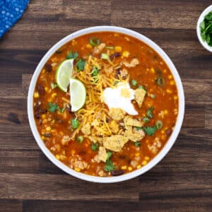 Taco soup topped with sour cream, tortilla chips, lime wedges, and fresh herbs, served in a white bowl.
