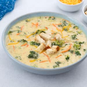 A bowl of broccoli cheddar soup on a white table, topped with crispy croutons. A blue towel, a cup of shredded cheddar cheese, and extra crusty bread sit nearby.