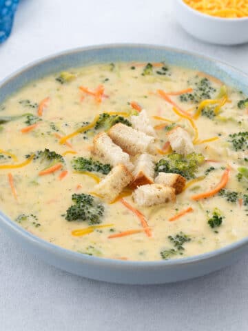 A bowl of broccoli cheddar soup on a white table, topped with crispy croutons. A blue towel, a cup of shredded cheddar cheese, and extra crusty bread sit nearby.