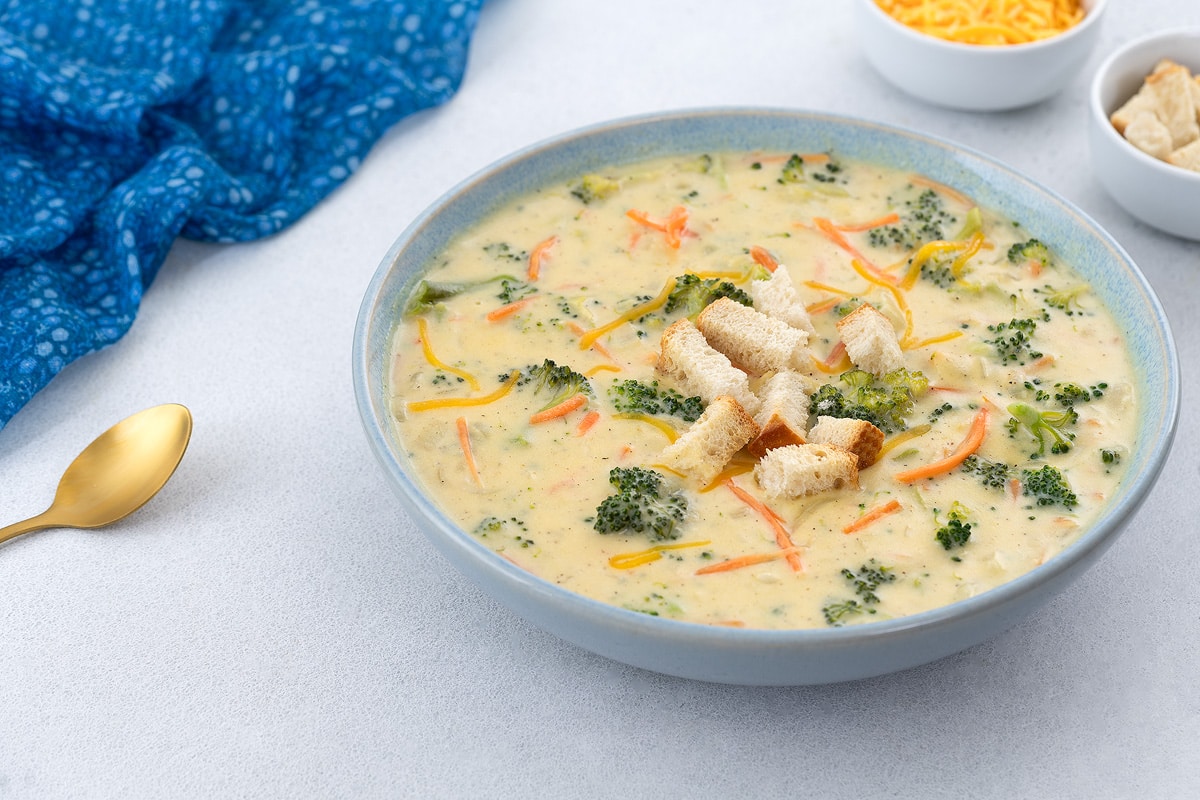 A bowl of broccoli cheddar soup on a white table, topped with crispy croutons. A blue towel, a gold spoon, a cup of shredded cheddar cheese, and extra crusty bread are placed nearby.