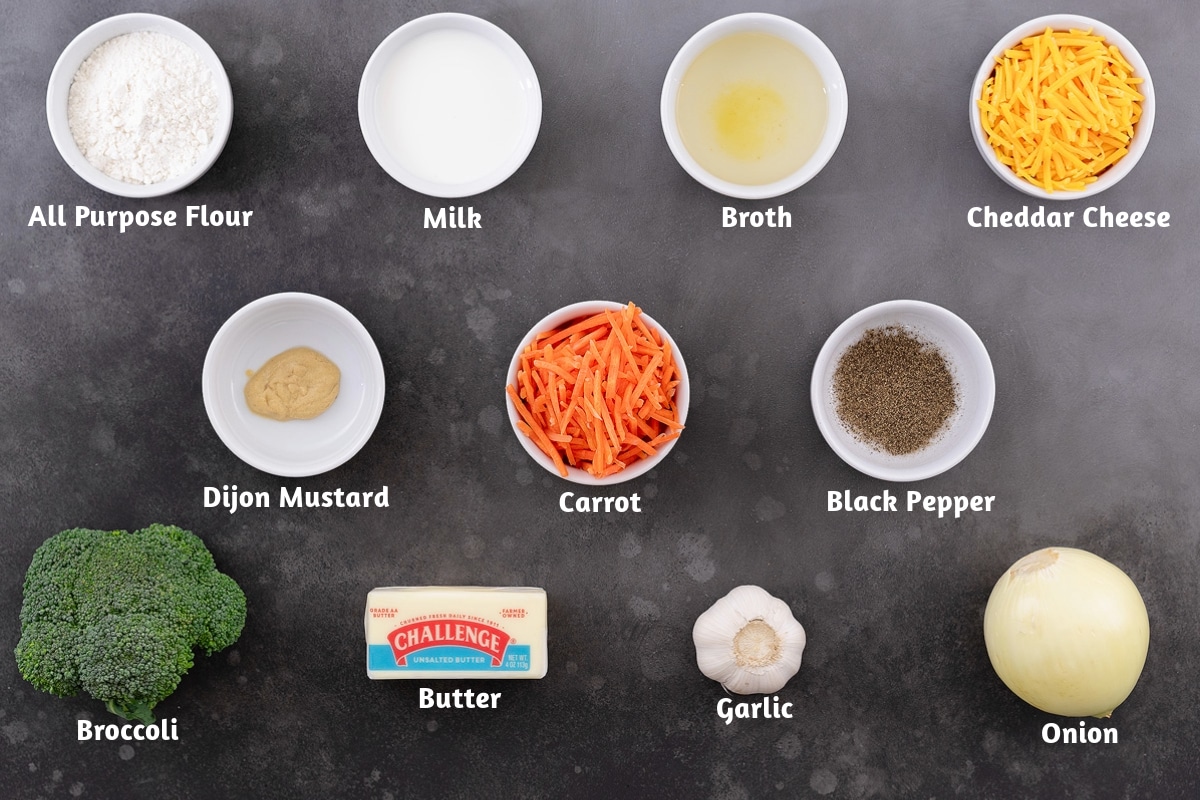 Broccoli cheddar soup ingredients arranged on a gray table, including all-purpose flour, milk, broth, cheddar cheese, Dijon mustard, carrot, black pepper, broccoli, butter, garlic, and onion.