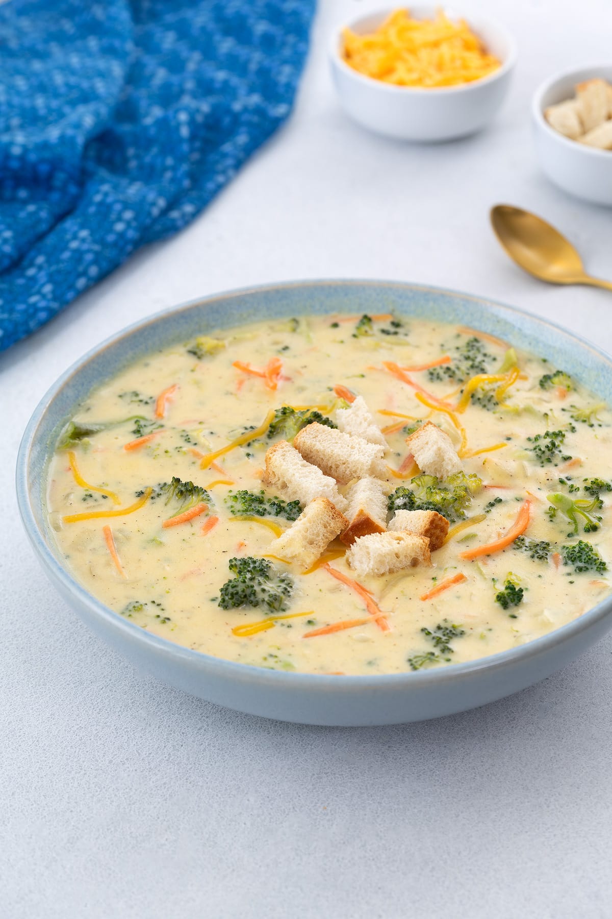 A bowl of broccoli cheddar soup on a white table, topped with crispy croutons. A blue towel, a gold spoon, a cup of shredded cheddar cheese, and extra crusty bread are placed nearby.
