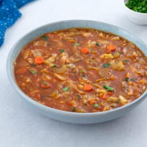 A bowl of cabbage soup placed on a white table, surrounded by a blue towel, and a cup of parsley.