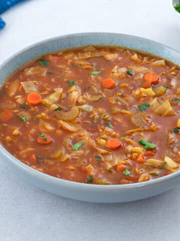 A bowl of cabbage soup placed on a white table, surrounded by a blue towel, and a cup of parsley.