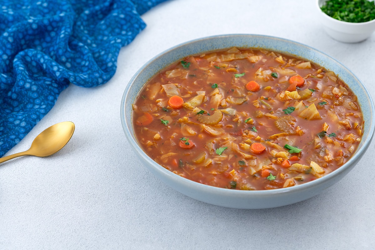 A bowl of cabbage soup placed on a white table, surrounded by a blue towel, a golden spoon, and a cup of parsley.