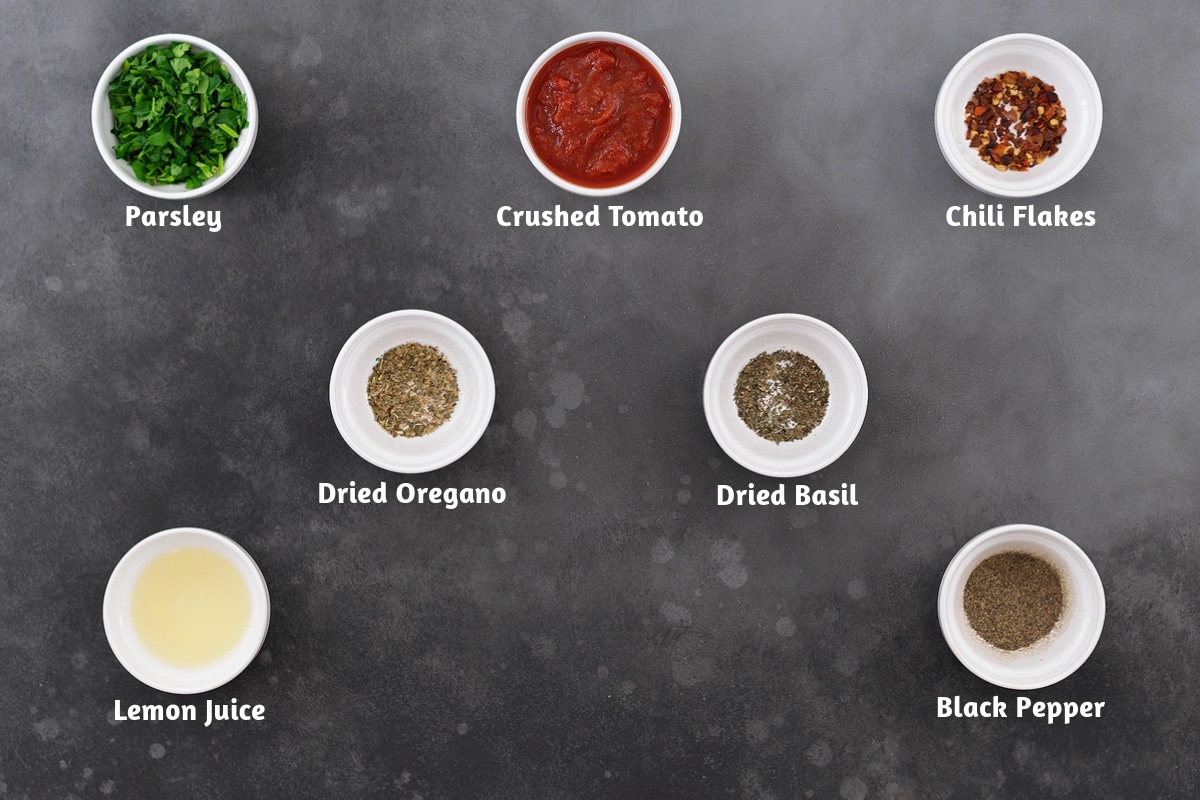 Cabbage soup recipe ingredients displayed on a gray table, featuring parsley, crushed tomatoes, chili flakes, dried oregano, dried basil, lemon juice, and black pepper.