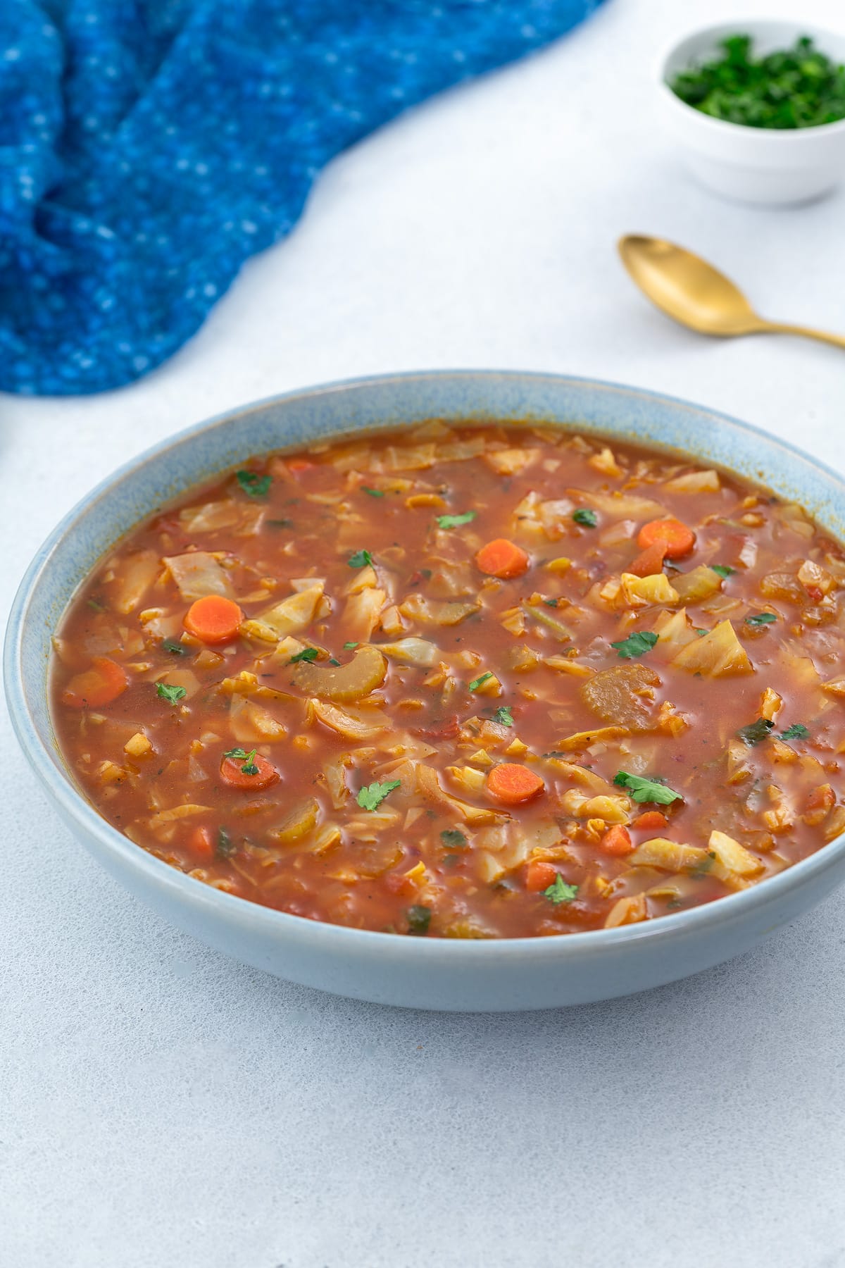A bowl of cabbage soup placed on a white table, surrounded by a blue towel, a golden spoon, and a cup of parsley.