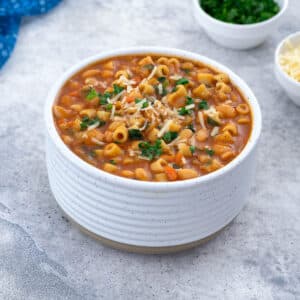 A white bowl filled with Pasta e Fagioli on a white table. Surrounding it are a blue towel, a small cup of parsley, and a cup of parmesan cheese.