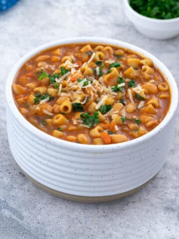 A white bowl filled with Pasta e Fagioli on a white table. Surrounding it are a blue towel, a small cup of parsley, and a cup of parmesan cheese.