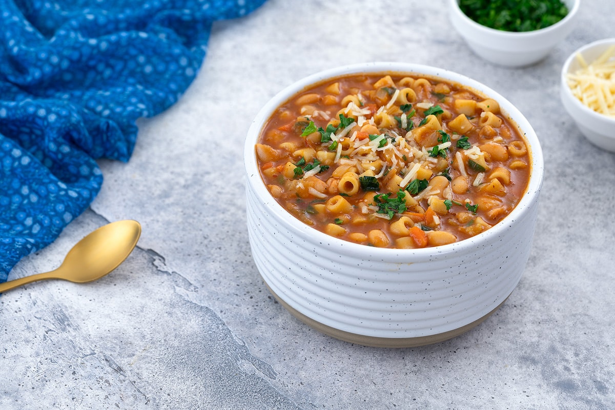 A white bowl filled with Pasta e Fagioli on a white table. Surrounding it are a blue towel, a golden spoon, a small cup of parsley, and a cup of parmesan cheese.
