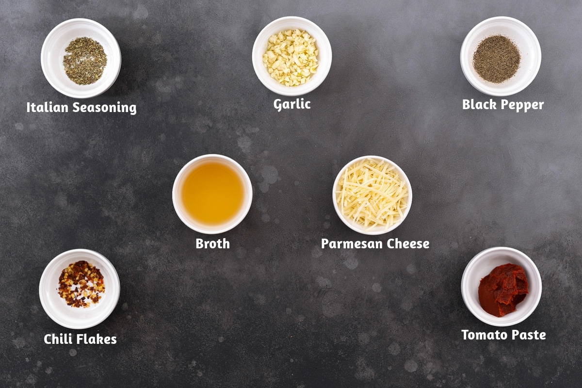 Pasta e Fagioli ingredients on a gray table, including Italian seasoning, garlic, black pepper, broth, parmesan cheese, chili flakes, and tomato paste.