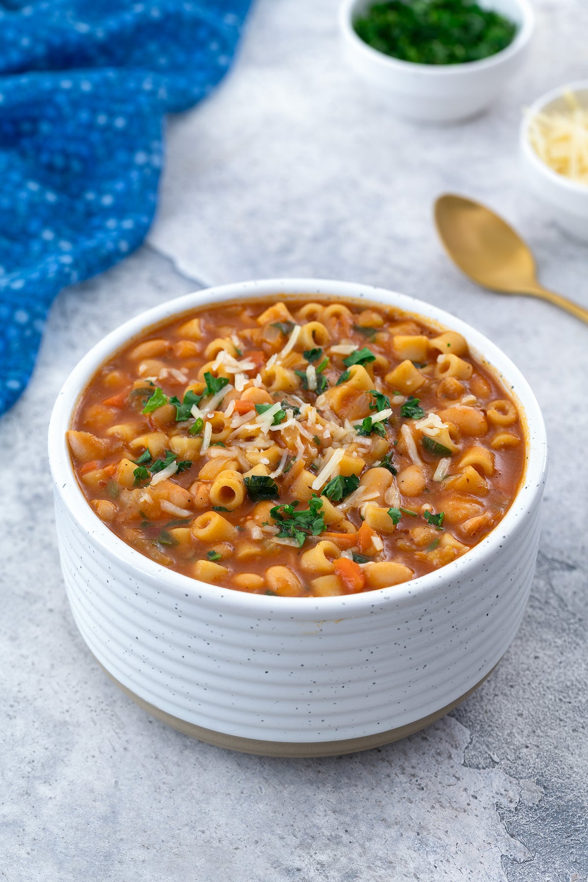 A white bowl filled with Pasta e Fagioli on a white table. Surrounding it are a blue towel, a golden spoon, a small cup of parsley, and a cup of parmesan cheese.