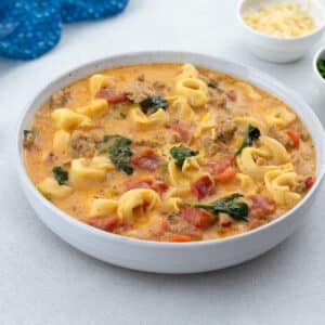 Homemade tortellini soup served in a white bowl on a white table. A blue towel, a cup of cheese, and fresh parsley leaves surround the bowl.