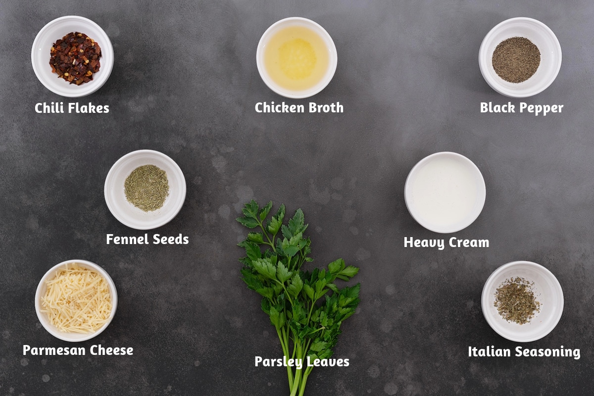 Tortellini soup ingredients arranged on a gray table. Chili flakes, chicken broth, and black pepper in one section; fennel seeds and heavy cream in another; and Parmesan cheese, parsley leaves, and Italian seasoning grouped together.