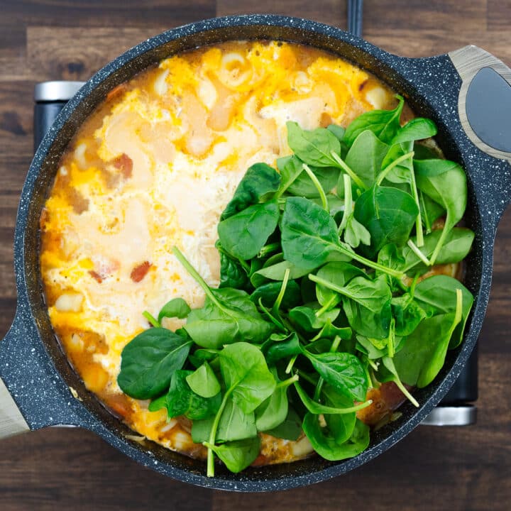 Pan with simmering tortellini soup, cream, parmesan cheese, and spinach leaves added.