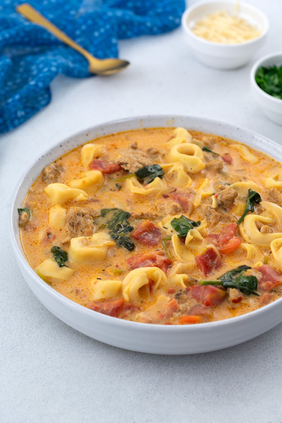 Homemade tortellini soup served in a white bowl on a white table. A blue towel, a golden spoon, a cup of cheese, and fresh parsley leaves surround the bowl.
