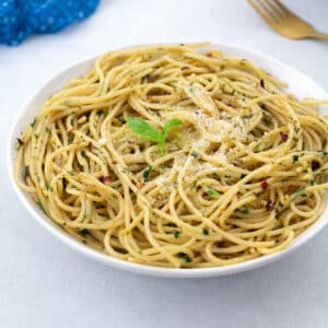 Homemade Spaghetti Aglio e Olio served in a white bowl, with a blue towel, a golden fork arranged around it.