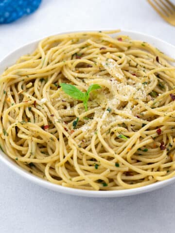 Homemade Spaghetti Aglio e Olio served in a white bowl, with a blue towel, a golden fork arranged around it.