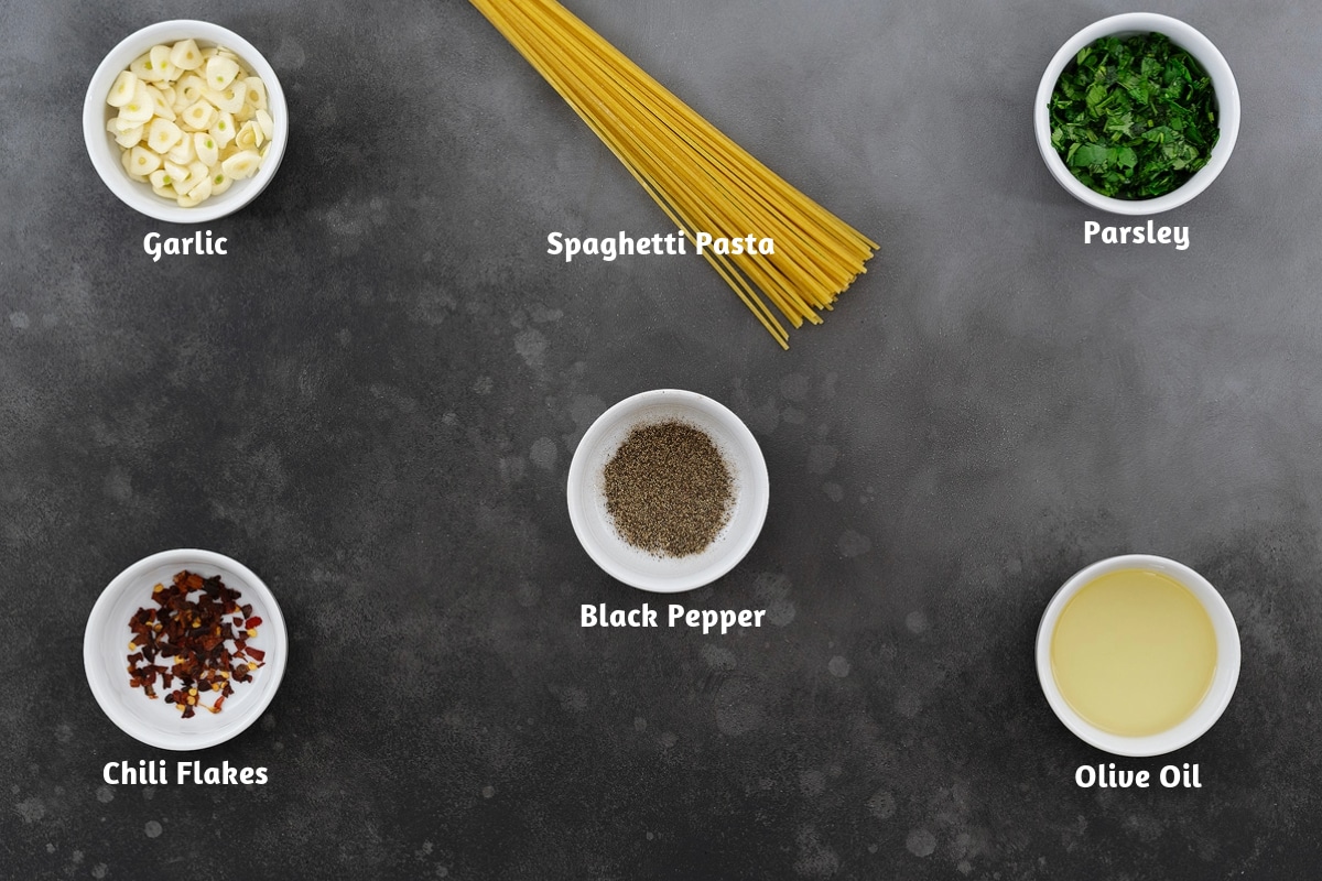 Spaghetti Aglio e Olio ingredients arranged on a gray table, including garlic, spaghetti pasta, parsley, black pepper, chili flakes, and olive oil.