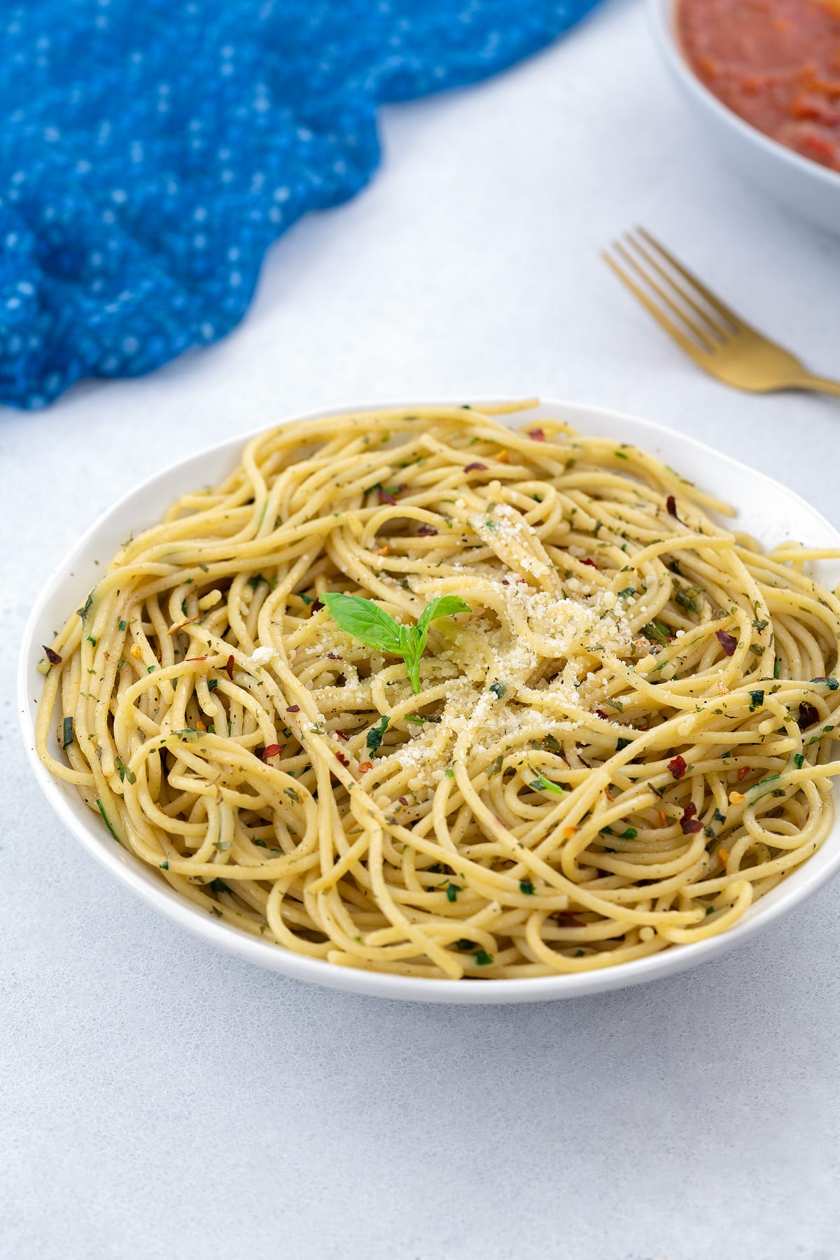 Homemade Spaghetti Aglio e Olio served in a white bowl, with a blue towel, a golden fork, and a bowl of chicken cacciatore arranged around it.