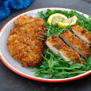 Whole and sliced chicken breast schnitzel on a white plate, served with baby arugula. A blue towel is placed beside the plate.
