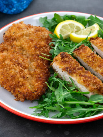 Whole and sliced chicken breast schnitzel on a white plate, served with baby arugula. A blue towel is placed beside the plate.