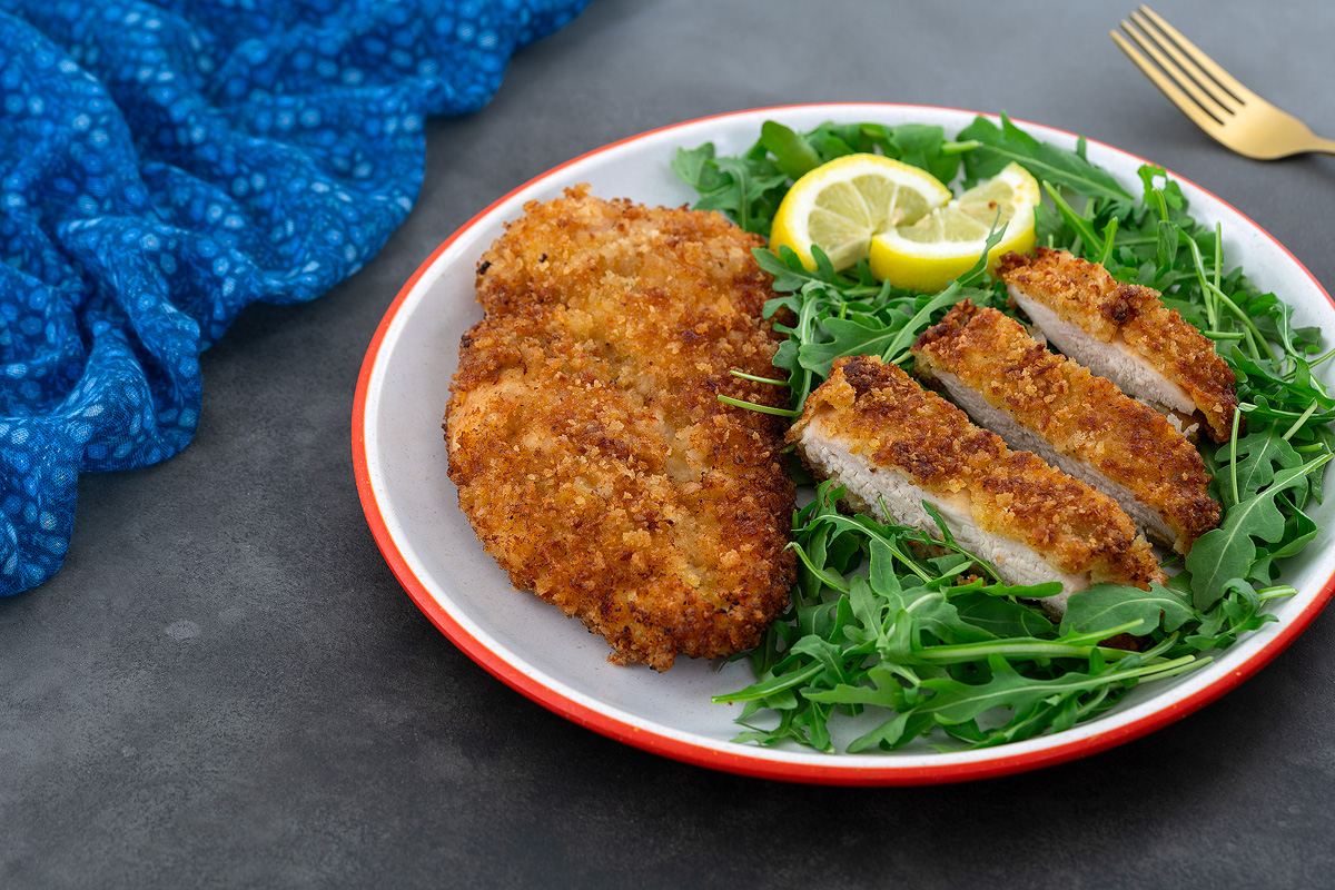 Whole and sliced chicken breast schnitzel on a white plate, served with baby arugula. A golden fork and blue towel are placed beside the plate.