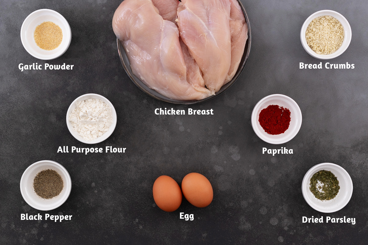 Chicken schnitzel ingredients arranged on a gray table, including chicken breast, garlic powder, bread crumbs, all-purpose flour, paprika, black pepper, egg, and dried parsley.