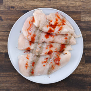A plate with boneless chicken breast seasoned with black pepper, paprika, and salt.