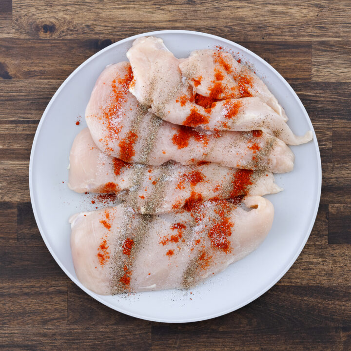 A plate with boneless chicken breast seasoned with black pepper, paprika, and salt.